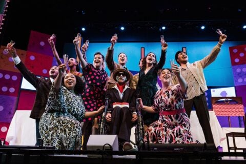 Group of people on stage raising glasses in a celebratory toast, with colorful set design in the background.