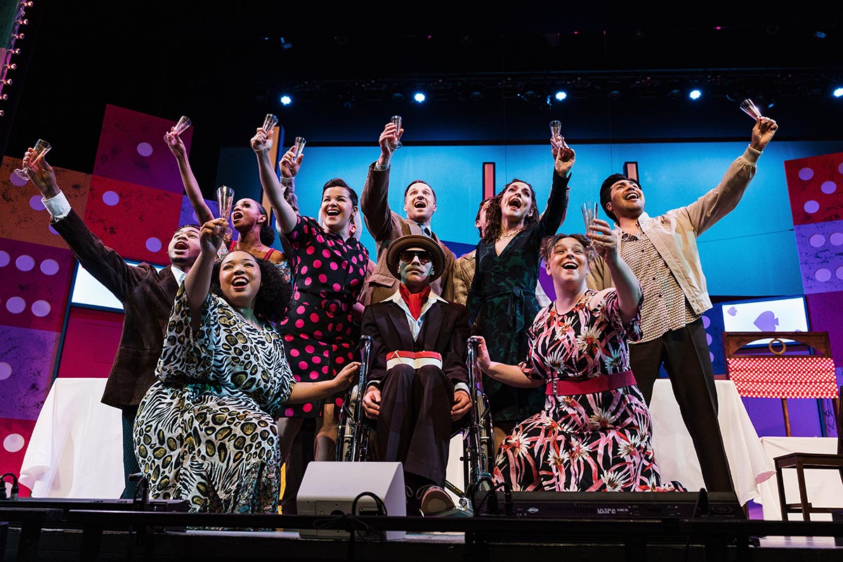 Group of people on stage raising glasses in a celebratory toast, with colorful set design in the background.