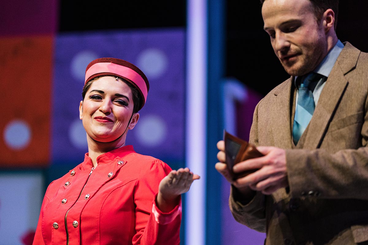 FLINT_Lucky Stiff 2026_0028 Two people on stage, one wearing a red uniform and hat gesturing with one hand, the other in a brown suit holding an open wallet.