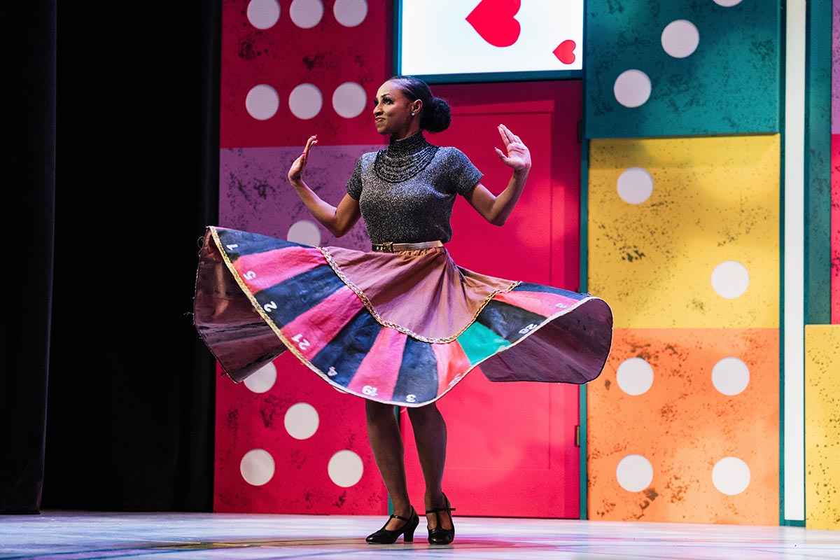 FLINT_Lucky Stiff 2026_0041 A dancer on stage wearing a colorful skirt resembling a roulette wheel, with large domino and playing card decorations in the background.
