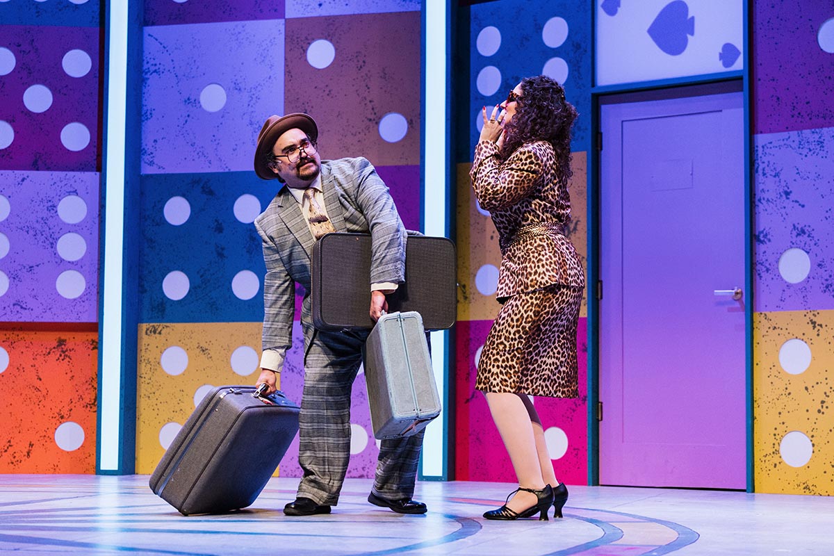 FLINT_Lucky Stiff 2026_0043 Two actors on stage with colorful domino-patterned walls; one carries three suitcases, the other wears a leopard print outfit.