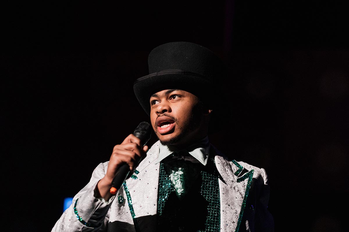 FLINT_Lucky Stiff 2026_0048 Person wearing a white and green sequined suit and a black top hat, holding a microphone against a dark background.