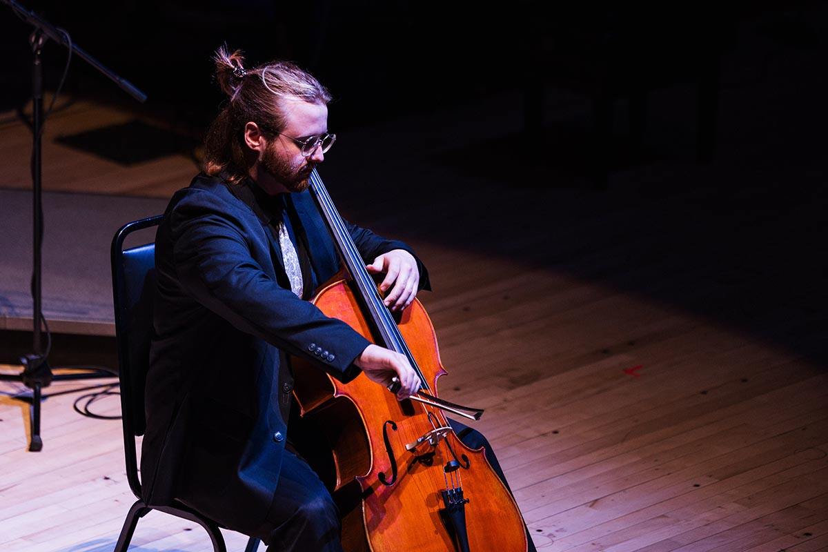 Person in a dark suit playing a cello while seated on a chair on a wooden stage.