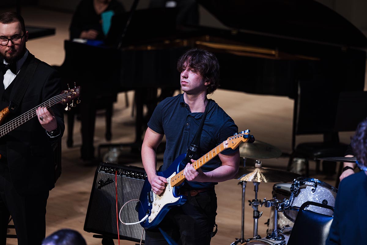 A musician playing an electric guitar on stage with a drum set and piano in the background.