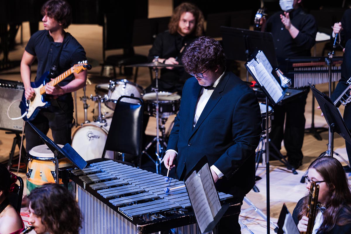 A group of musicians performing on stage, including a vibraphone player in a suit, a guitarist, a drummer, and other instrumentalists.