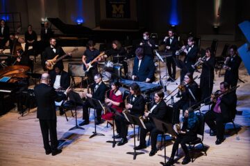 Orchestra performing on stage with conductor leading musicians playing various instruments including saxophones, trumpets, guitars, drums, and piano.