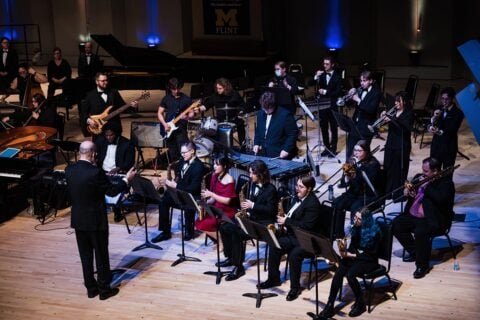Orchestra performing on stage with conductor leading musicians playing various instruments including saxophones, trumpets, guitars, drums, and piano.