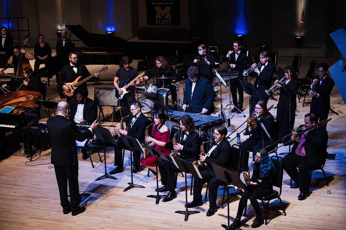 Orchestra performing on stage with conductor leading musicians playing various instruments including saxophones, trumpets, guitars, drums, and piano.