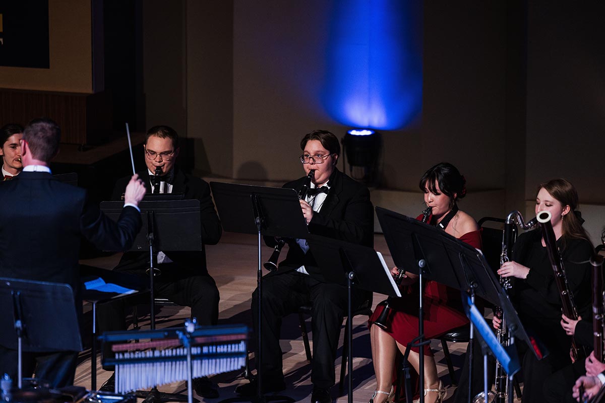 Orchestra conductor leading musicians playing woodwind instruments on stage with blue spotlight in the background.