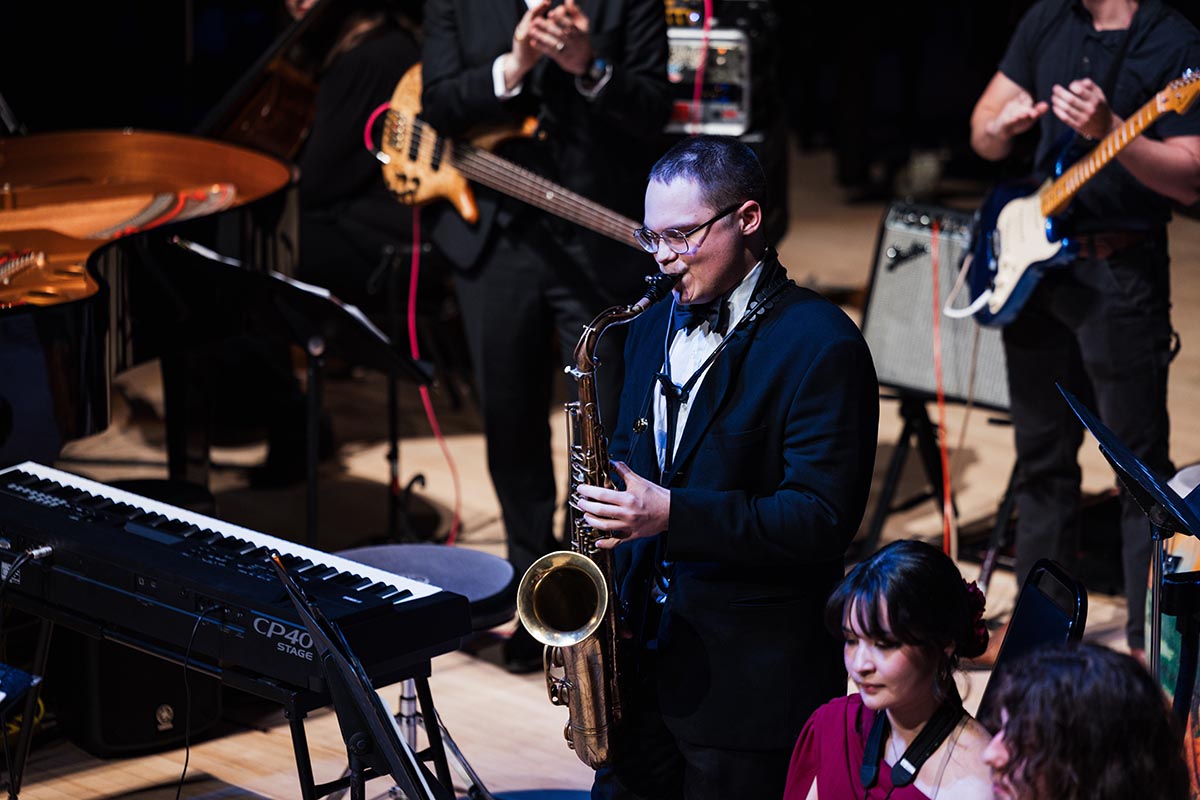 A musician in a suit playing a saxophone on stage with other musicians and instruments, including a keyboard and guitars.