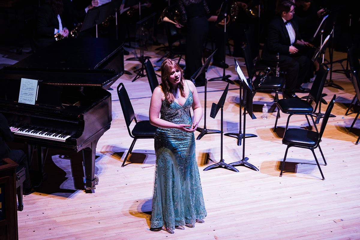 A woman in a sparkling teal gown stands on a stage in front of a grand piano and empty chairs with music stands, performing with an orchestra.