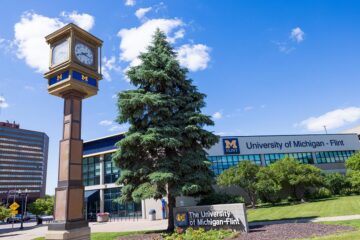 Clock tower and sign in front of University of Michigan-Flint campus building on a sunny day.