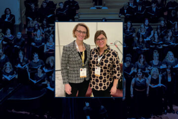 Two women wearing conference badges stand side by side indoors. The background is a choral performance.