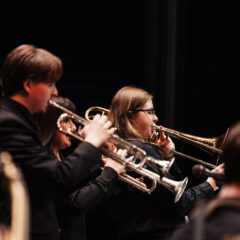 A group of musicians playing brass instruments, including trumpets and a trombone, on stage in a dark setting.
