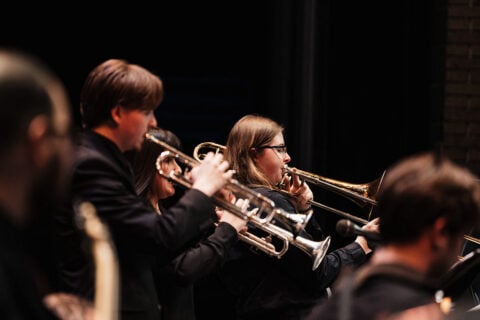 A group of musicians playing brass instruments, including trumpets and a trombone, on stage in a dark setting.