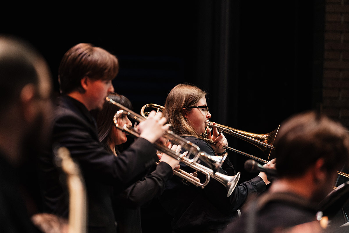 A group of musicians playing brass instruments, including trumpets and a trombone, on stage in a dark setting.