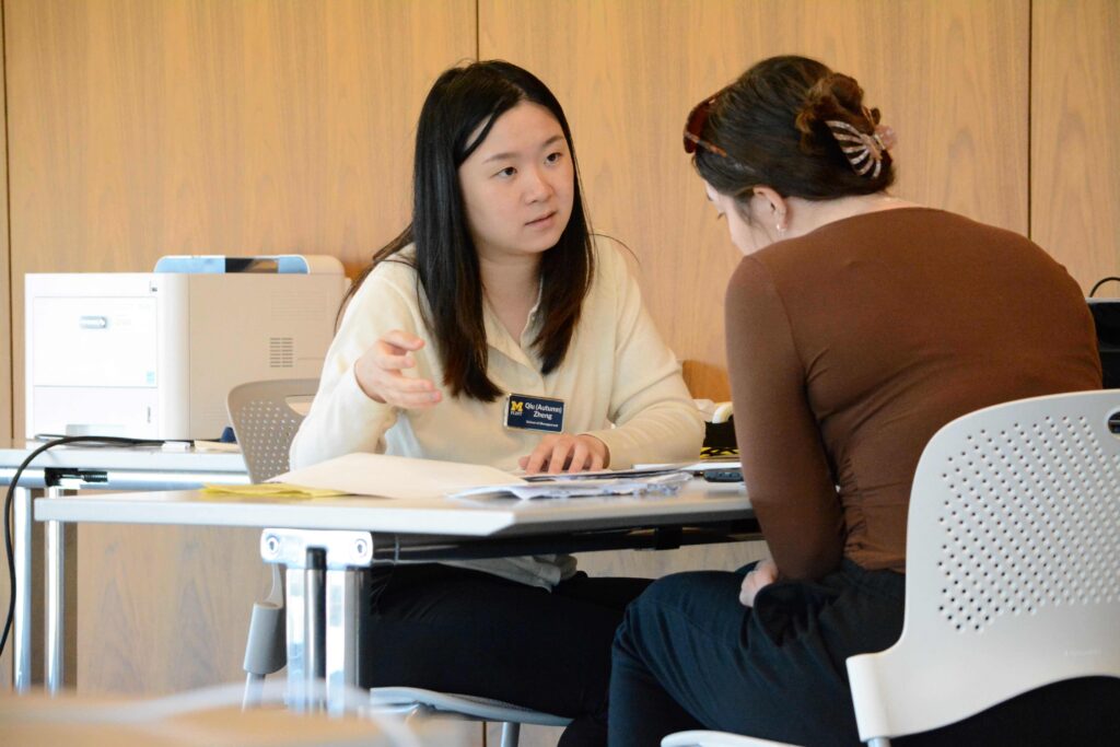A woman wearing a University of Michigan-Flint name tag sits at a white table gesturing with one hand as she speaks to a young woman about tax documents.
