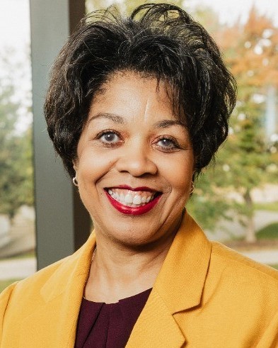 A professional portrait of a woman with short, dark hair and a bright smile. She is wearing a mustard-yellow blazer over a maroon blouse and small hoop earrings. The background features a window overlooking blurred green and orange trees.