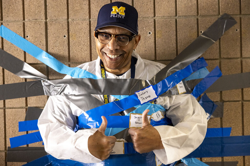 Chancellor Laurence Alexander smiling and giving a thumbs up while being duct taped to a brick wall.