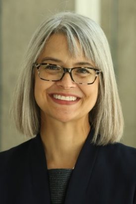 Person with straight, shoulder-length gray hair wearing a dark blazer and a dark top.