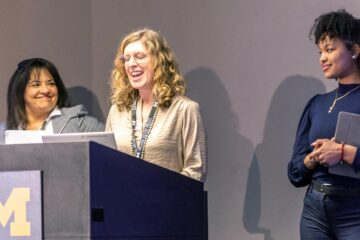 Three women standing at a podium with a University of Michigan logo, one speaking and the others listening. Photo by Annaleigh Barker.