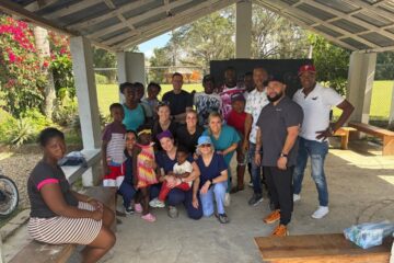 Group of people, including children and adults from the community, gathered with the medical mission team on final clinic day under a covered outdoor area with benches and greenery in the background.