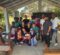 Group of people, including children and adults from the community, gathered with the medical mission team on final clinic day under a covered outdoor area with benches and greenery in the background.