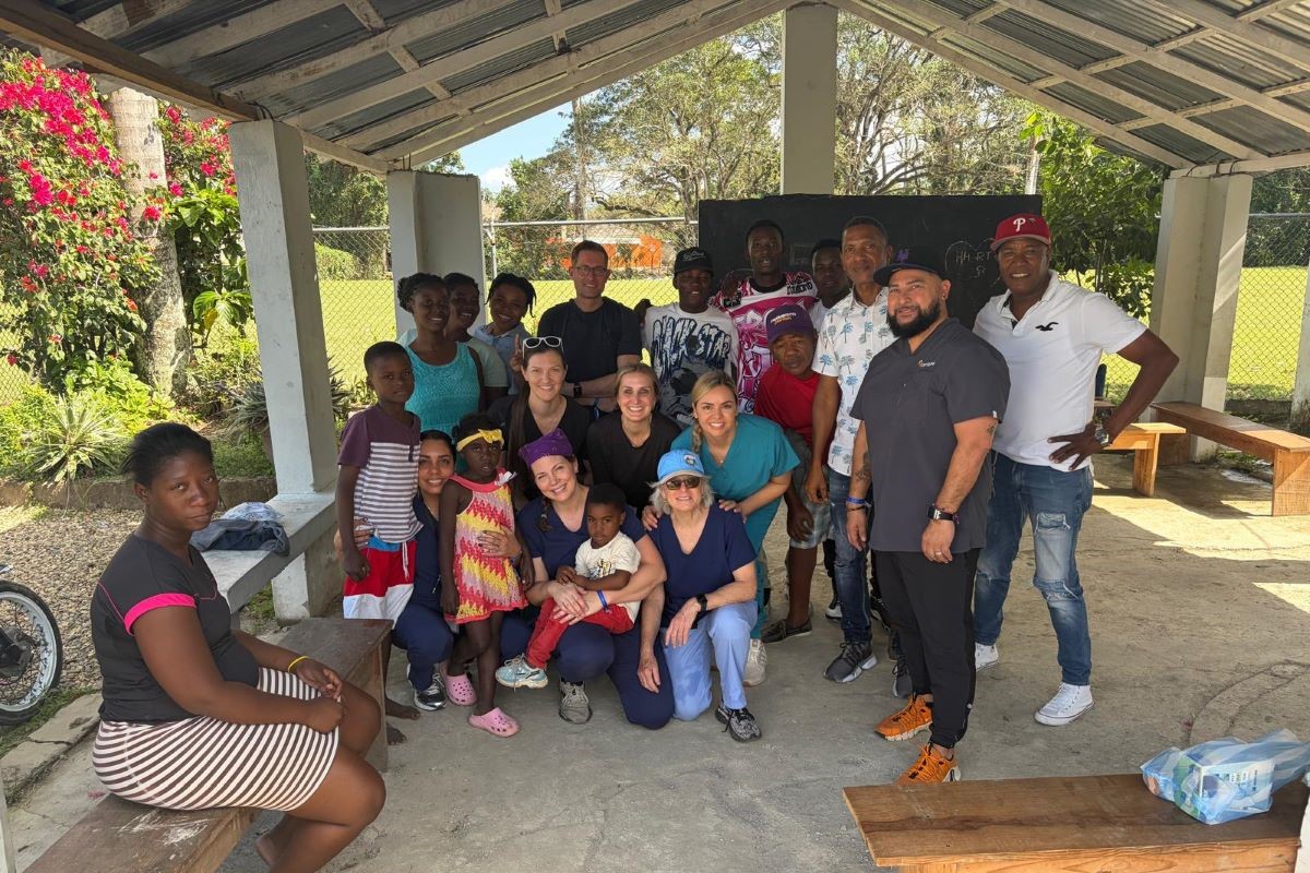 Group of people, including children and adults from the community, gathered with the medical mission team on final clinic day under a covered outdoor area with benches and greenery in the background.
