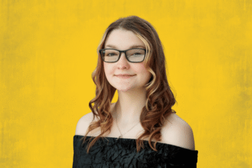 Person with long curly hair wearing an off-shoulder black top against a yellow background.