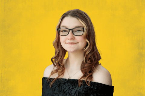 Person with long curly hair wearing an off-shoulder black top against a yellow background.