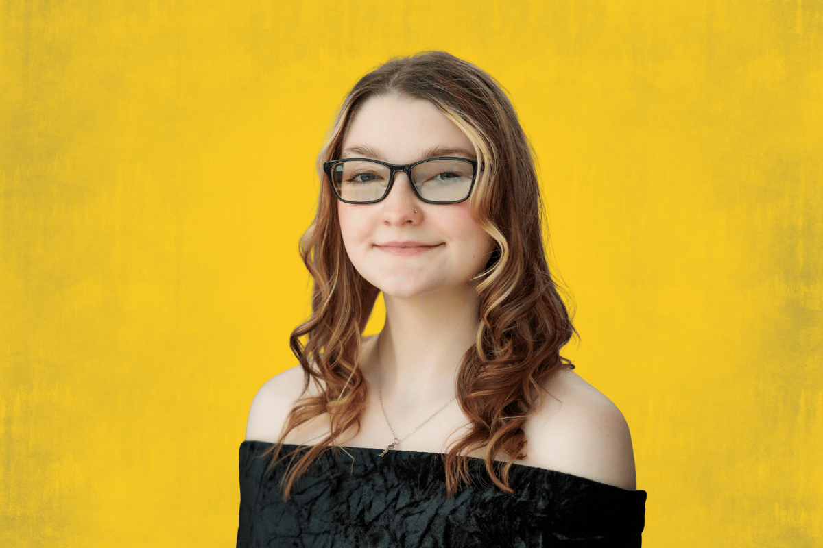Person with long curly hair wearing an off-shoulder black top against a yellow background.