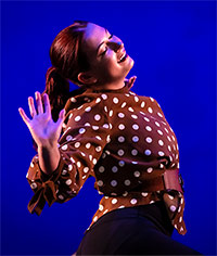 Person in a brown polka dot shirt with a belt, posing with one hand raised against a blue background.