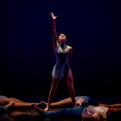 A dancer standing center stage with one arm raised, surrounded by other dancers lying on the floor in a dark setting.
