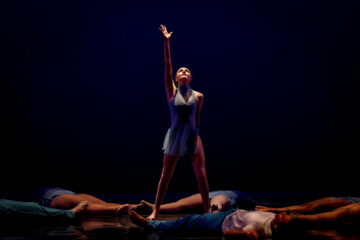 A dancer standing center stage with one arm raised, surrounded by other dancers lying on the floor in a dark setting.