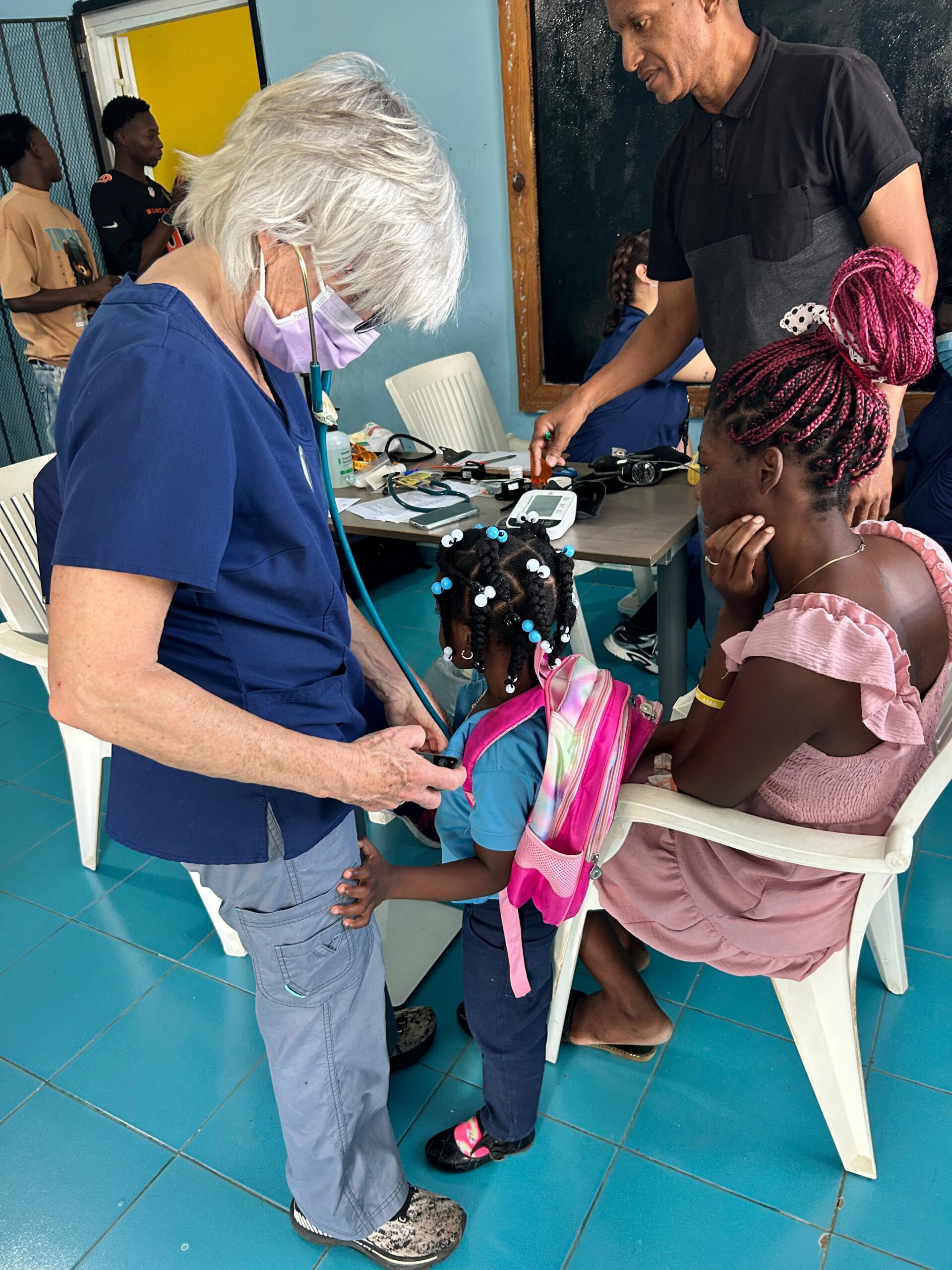 A health care worker wearing scrubs and a mask uses a stethoscope to examine a young child with a pink backpack, while a woman and a man are nearby in a clinic setting.