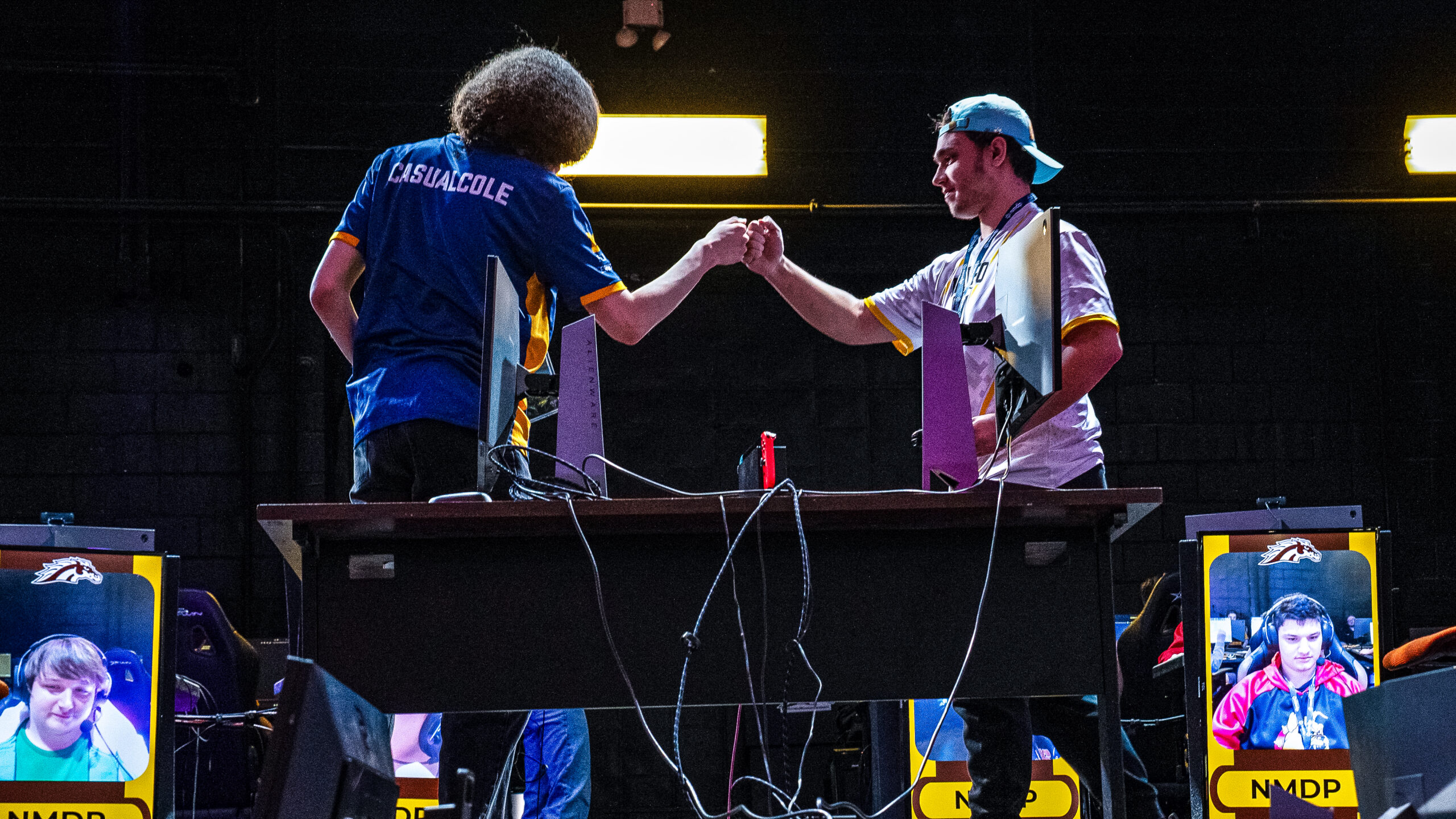 Two esports players fist bump over a desk with gaming monitors during a competition.