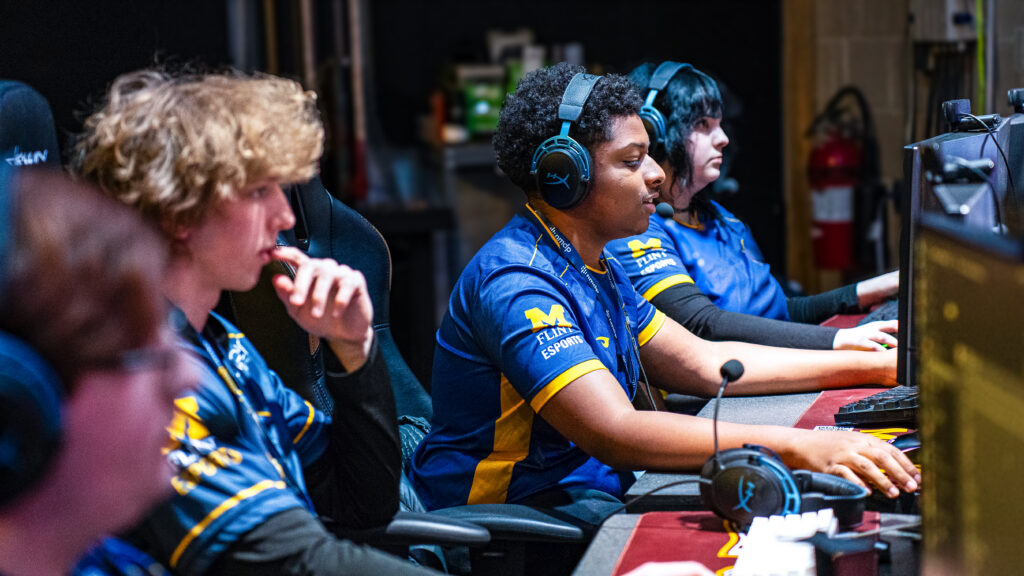 A group of esports players wearing blue and yellow Michigan Flint Esports jerseys and headsets, sitting in front of computer monitors and gaming equipment.
