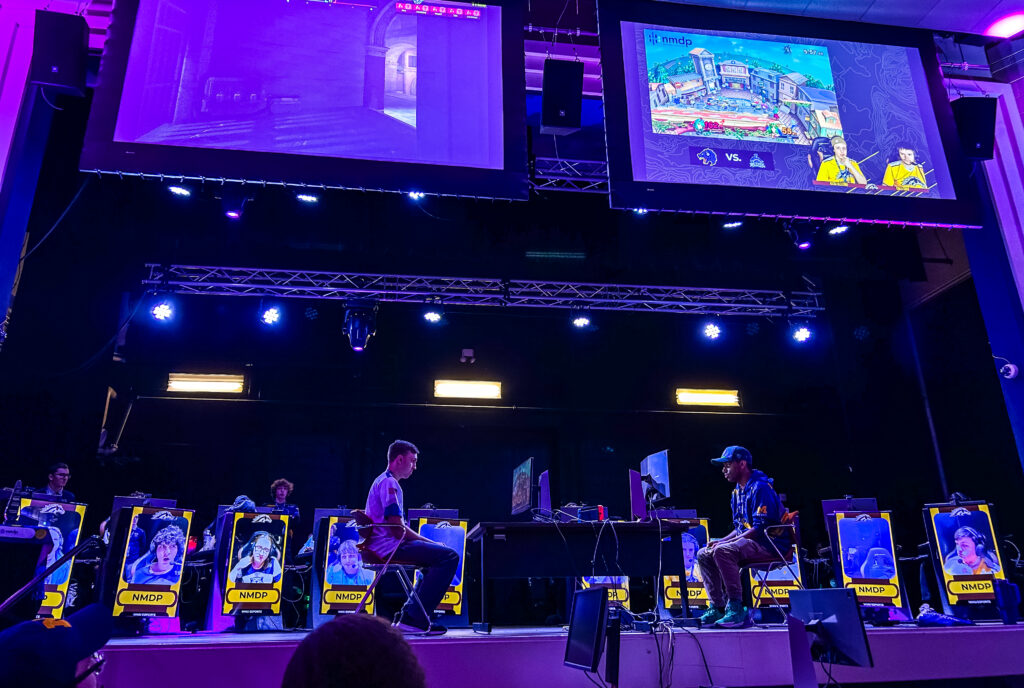 Two players seated on stage facing each other with monitors, large screens above showing gameplay from two different video games. 