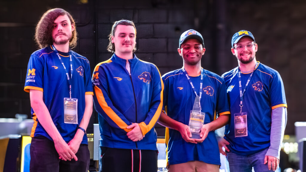 Four esports team members wearing matching blue and yellow jerseys with lanyards, one holding a trophy.