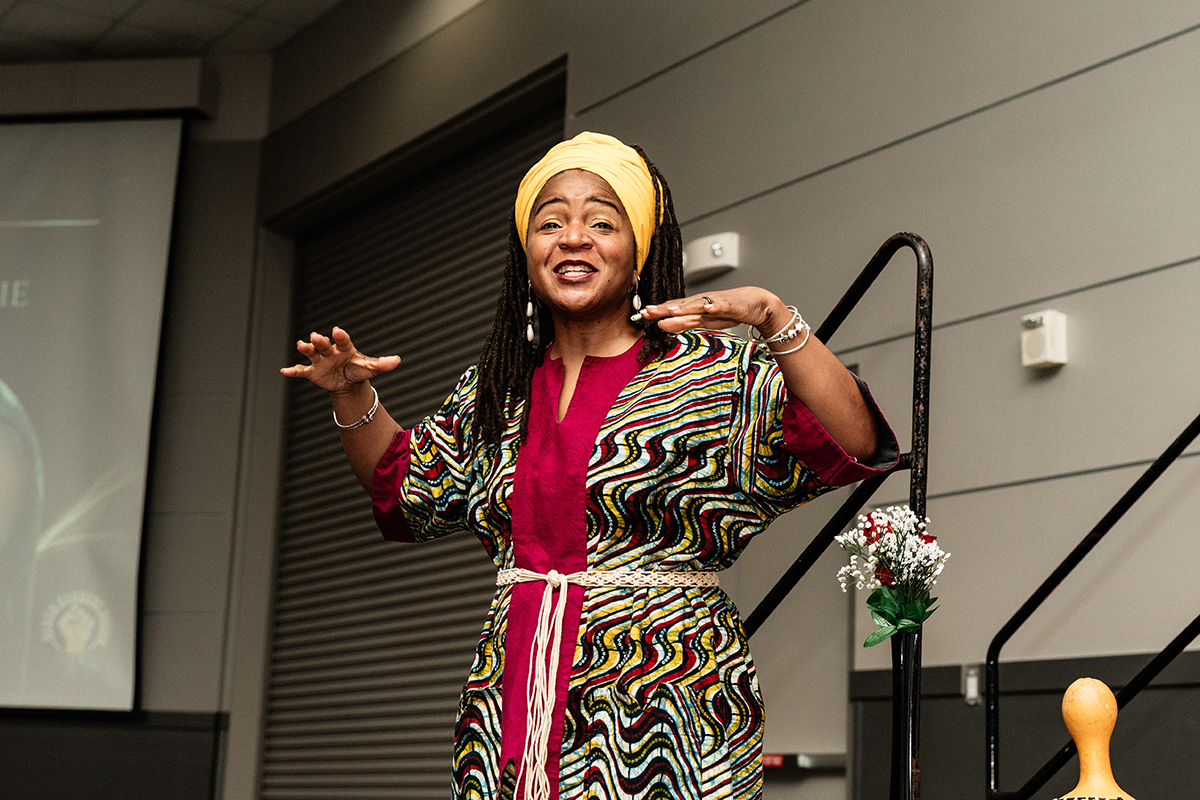 FLINT_BLAK Graduation Celebration 2026_0014 A person wearing a colorful patterned dress and a yellow headwrap is speaking or gesturing with both hands raised indoors near a staircase and a small vase of flowers.