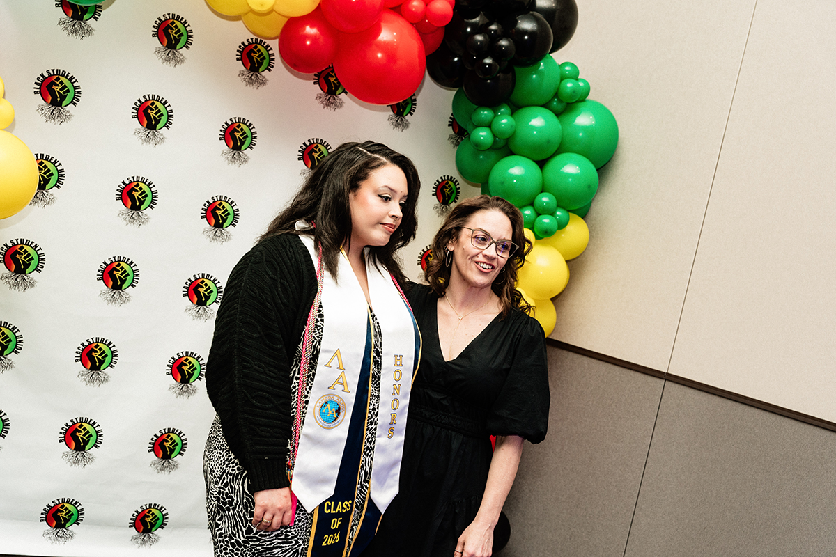 FLINT_BLAK Graduation Celebration 2026_0041 Two women standing in front of a Black Student Union backdrop with red, yellow, green, and black balloons; one woman wears a white honors sash.