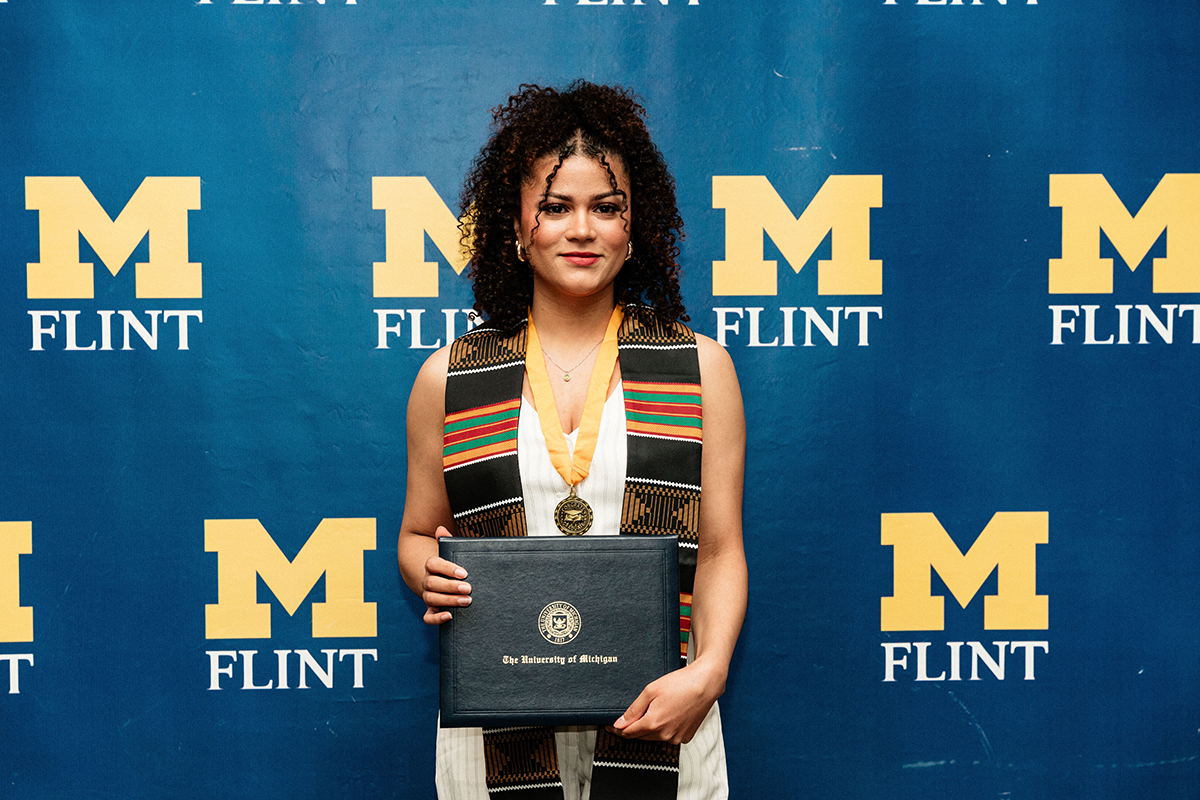 FLINT_BLAK Graduation Celebration 2026_0055 Person wearing a medal and a colorful stole holding a University of Michigan diploma in front of a blue backdrop with yellow "M Flint" logos.