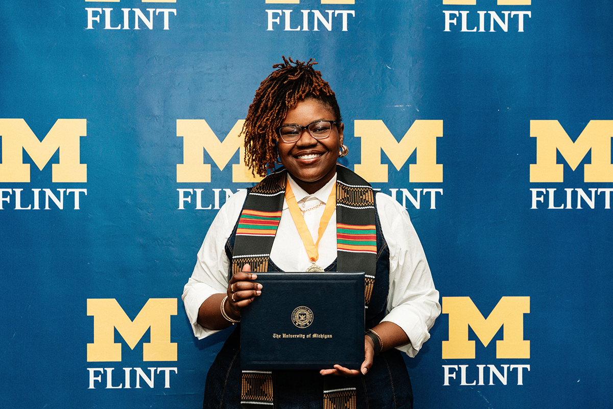 FLINT_BLAK Graduation Celebration 2026_0056 Person holding a University of Michigan diploma in front of a blue backdrop with yellow "M Flint" logos.