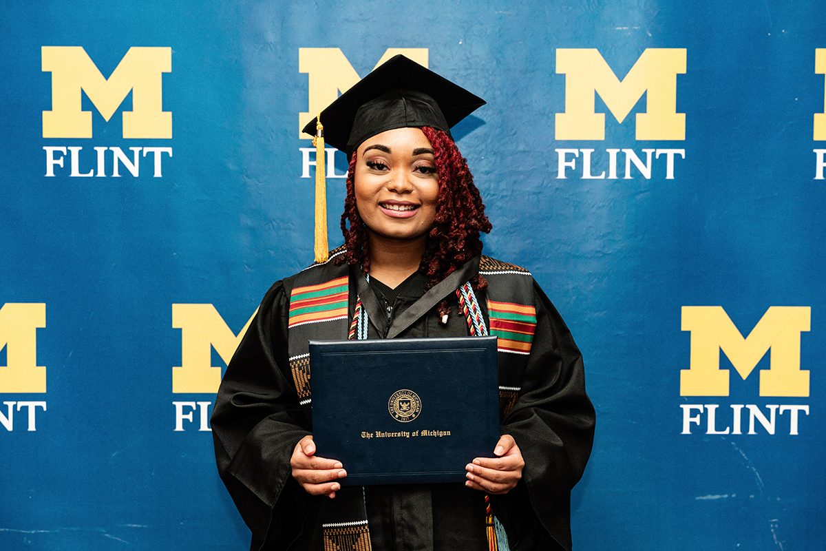 FLINT_BLAK Graduation Celebration 2026_0057 Graduate in cap and gown holding a University of Michigan diploma in front of a blue backdrop with multiple yellow "M Flint" logos.