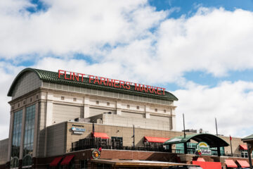 Flint Farmers' Market building with a large red sign on the roof and a partly cloudy sky above.
