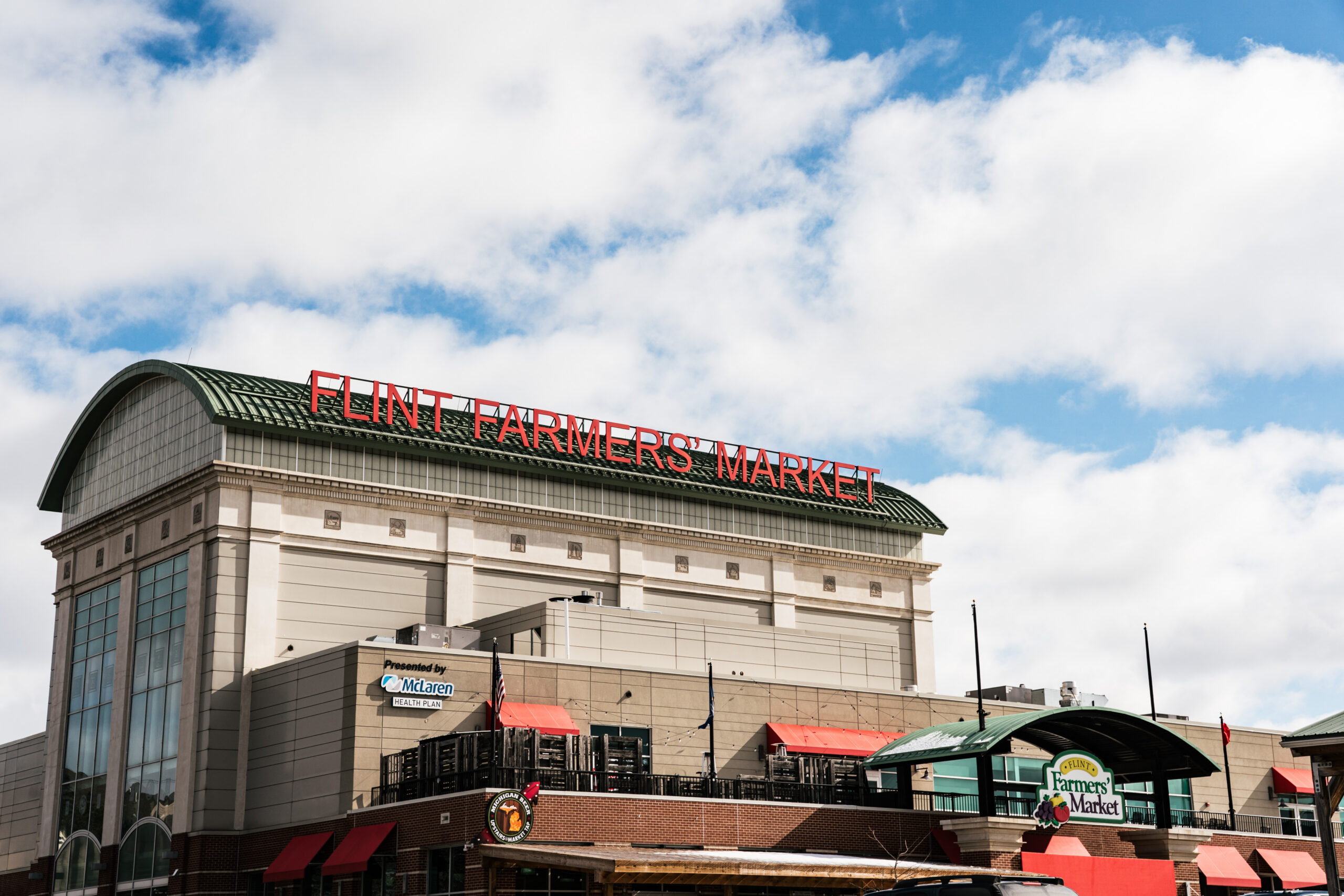 Flint Farmers' Market building with a large red sign on the roof and a partly cloudy sky above.