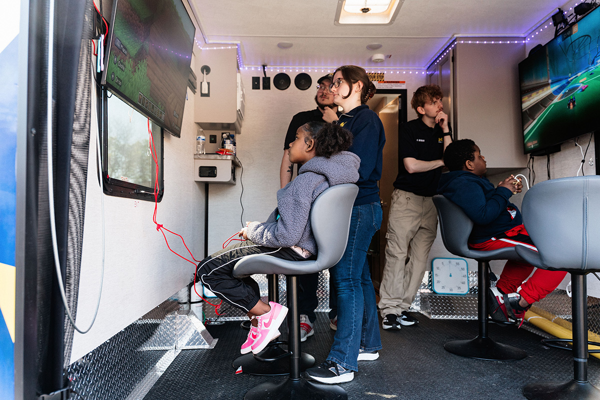 Children and adults playing video games on two wall-mounted screens inside a gaming trailer.