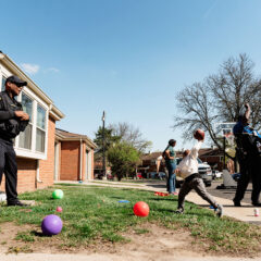 Police officers and children playing basketball and catch outside in a residential neighborhood on a sunny day.