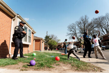 Police officers and children playing basketball and catch outside in a residential neighborhood on a sunny day.