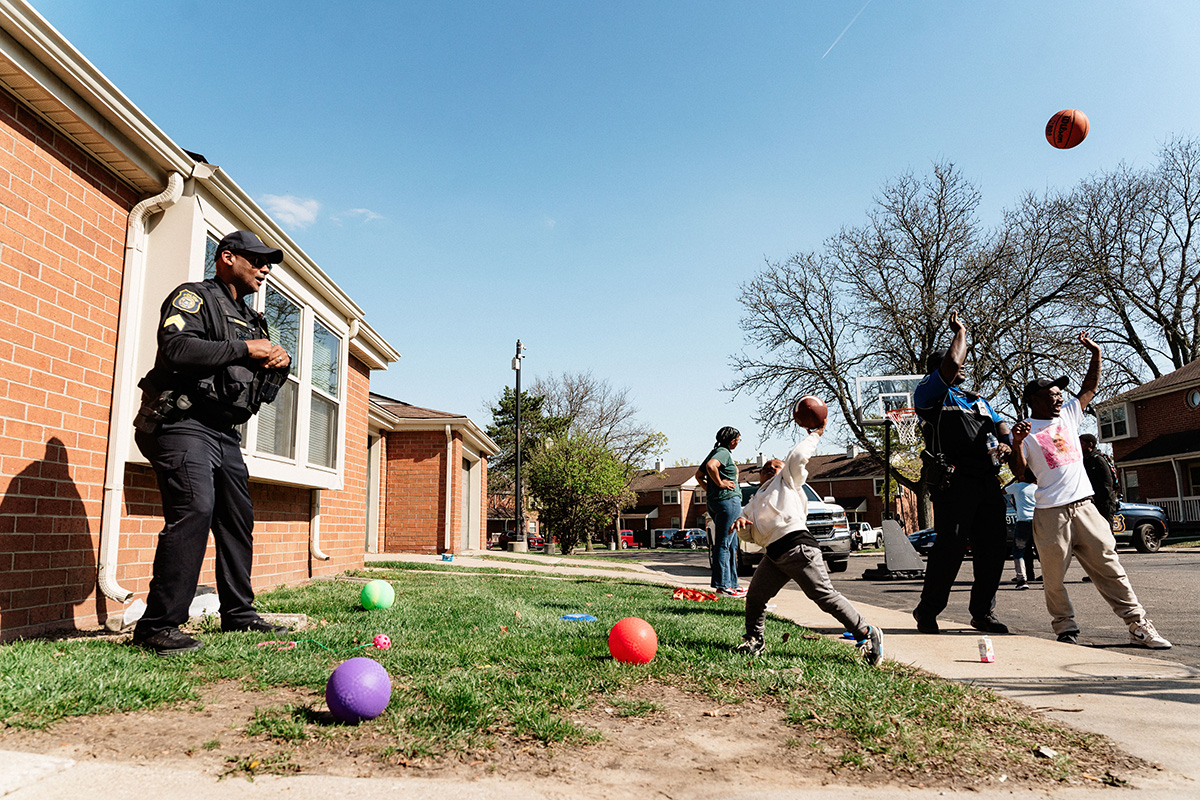 Police officers and children playing basketball and catch outside in a residential neighborhood on a sunny day.